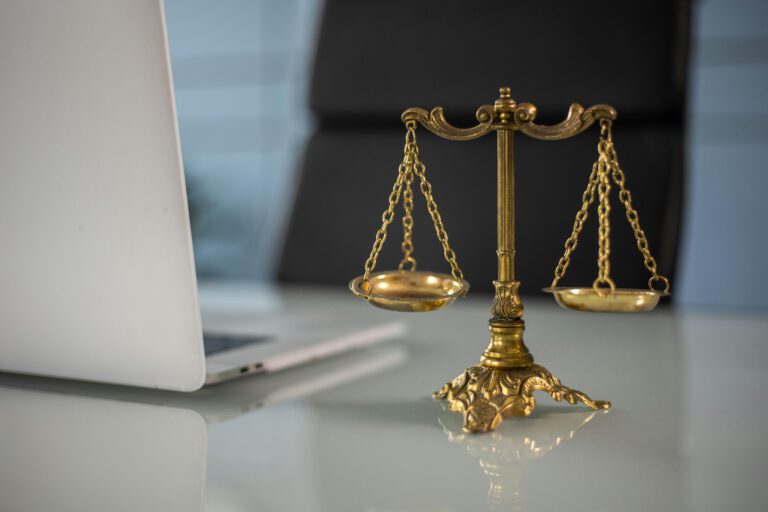 A brass balance scale placed on a glass desk next to an open silver laptop, symbolizing justice and legal work in a modern office setting.