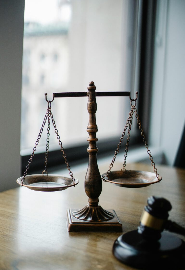 A bronze balance scale sits on a wooden desk near a window, symbolizing justice. Beside it, a wooden judge's gavel rests on the surface. The background shows an out-of-focus cityscape through the glass.