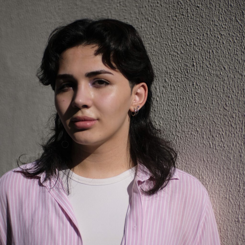 Alex with shoulder-long black hair, wearing a pink shirt looking into the camera