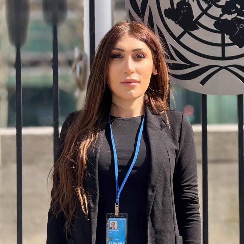 Lilith with long brown haid wearing a black suit jacket and black shirt looking into the camera