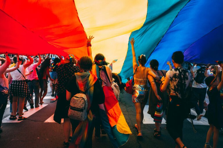People standing in the street on a crosswalk under a huge rainbow flag
