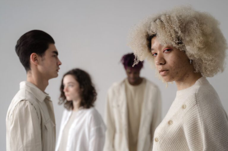 A woman with voluminous curls looks directly at the camera, while three softly focused people in neutral clothing stand behind her in a calm, contemplative studio portrait.